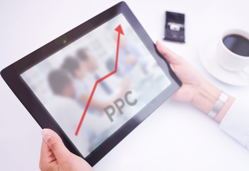 an image of tablet screen showing a line graph starting from the bottom and going up (in Red) showing the performance of an Adwords campaign against a blurred out image of four consultants sat around a table with the words PPC positioned middle bottom