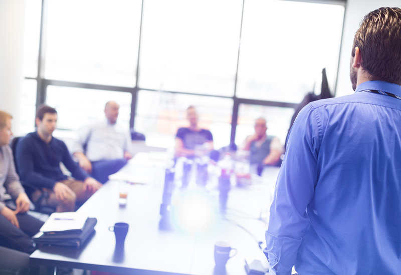an image showing a public relations consultant standing up and giving a presentation talk to a number of employees sitting around a table